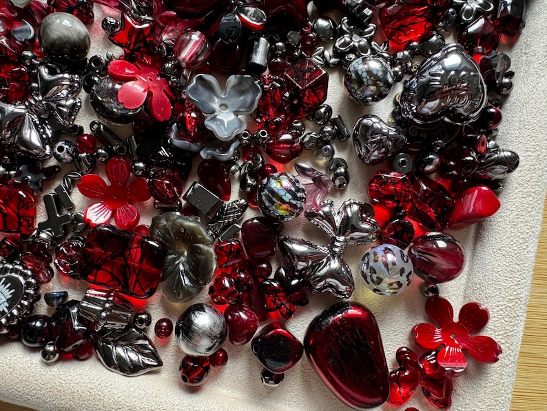 May include: A collection of assorted craft beads in various shapes and sizes, featuring a color palette of deep red, metallic silver, and charcoal gray. The assortment includes flower-shaped beads, heart-shaped charms, spheres, and faceted pieces.
