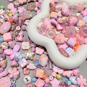 Pode incluir: Uma cole&ccedil;&atilde;o de contas e berloques em cores pastel em v&aacute;rias formas, incluindo estrelas, flores e cubos. As contas est&atilde;o espalhadas sobre uma superf&iacute;cie cinza e tamb&eacute;m em uma tigela branca com bordas onduladas. As cores incluem rosa, roxo, laranja e transparente.