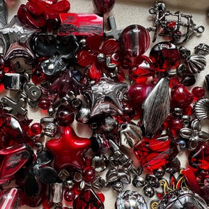 May include: A collection of assorted craft beads and charms in shades of red, black, and metallic silver. The assortment includes star-shaped beads, floral-shaped pieces, spherical beads, and various geometric shapes with different textures and finishes.