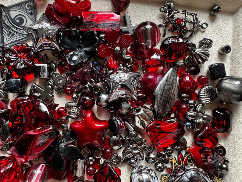 May include: A collection of assorted craft beads and charms in shades of red, black, and metallic silver. The assortment includes star-shaped beads, floral-shaped pieces, spherical beads, and various geometric shapes with different textures and finishes.