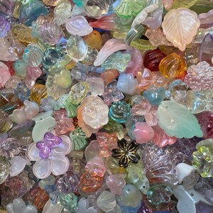 May include: A large collection of assorted translucent and opaque beads in various shapes including flowers, leaves, and butterflies. The beads feature a spectrum of colors such as pink, blue, yellow, green, and purple with iridescent finishes.