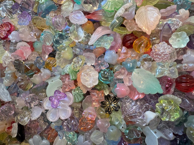 May include: A large collection of assorted translucent and opaque beads in various shapes including flowers, leaves, and butterflies. The beads feature a spectrum of colors such as pink, blue, yellow, green, and purple with iridescent finishes.