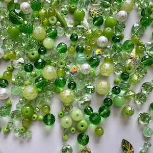 Op de afbeelding: Een verzameling losse kralen in diverse tinten groen, waaronder limoen, smaragd en olijf. Het assortiment bevat ronde, gefacetteerde en gladde glazen en plastic kralen, sommige met iriserende afwerkingen, verspreid over een effen oppervlak.
