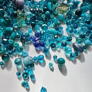 May include: A selection of blue and teal beads in various shapes and sizes, including faceted, round, and irregular forms. The beads are arranged on a white surface, with shadows visible. The collection includes glass and other materials.