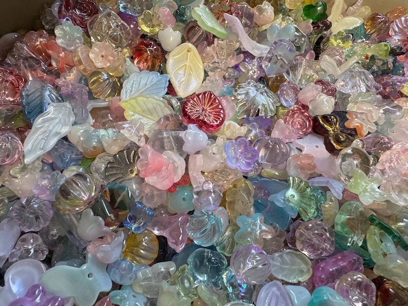May include: A large collection of assorted translucent acrylic beads in various shapes including flowers, leaves, and hearts. The beads feature a pastel color palette with pink, blue, yellow, green, and clear tones, each with a smooth, glossy finish.
