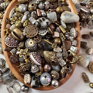 May include: A wooden bowl overflowing with a collection of beads and charms in diverse shapes, sizes, and colours. The beads display metallic gold, bronze, and silver hues, some with detailed patterns. Clear and iridescent beads are also present.