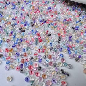 May include: A large collection of small, round, faceted glass beads in a variety of translucent colours including clear, pink, blue, purple, and yellow. Each bead features a central hole and a reflective, shimmering surface texture.
