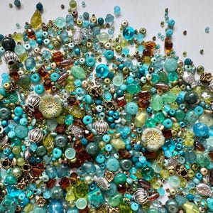 Peut inclure: Un assortiment de perles de différentes formes et tailles, dans des tons turquoise, vert, marron et argent. Certaines perles ont une finition métallique et des motifs floraux.