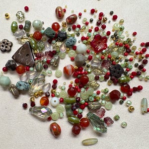 May include: A collection of assorted loose beads in various shapes, sizes, and textures. The mix includes red, green, clear, and brown glass, stone, and plastic beads, featuring floral-shaped pieces, faceted crystals, and smooth round spheres.