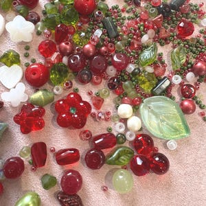 May include: A collection of loose craft beads in various shades of red, green, and white. The assortment includes round, faceted, and leaf-shaped beads, along with flower and heart-shaped pieces in glass, plastic, and opaque finishes of various sizes.