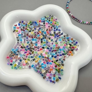 Puede incluir: Un plato blanco en forma de flor lleno de una variedad de pequeñas cuentas de colores. También hay una pulsera de cuentas multicolores. Las cuentas son en tonos de rosa, azul, verde, amarillo y morado. El fondo es gris liso.