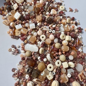 May include: A selection of beads in a palette of browns, beiges, and pinks. The mix includes round, square, and organic-shaped beads, some with metallic details. The beads are spread out, indicating a craft supply or jewellery-making theme.