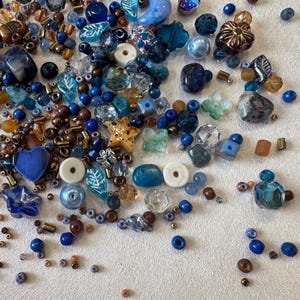 May include: A collection of loose craft beads in various shades of blue, brown, and clear glass. The assortment includes star-shaped, heart-shaped, leaf-shaped, and round beads, along with small metallic spacers and tiny seed beads in mixed sizes.