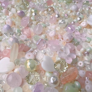 May include: A collection of assorted craft beads in various shades of pastel pink, soft green, white, and translucent clear. The mix includes faceted crystals, smooth round pearls, heart-shaped beads, and flower-shaped pieces of varying sizes.