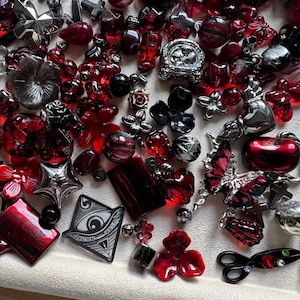 May include: A collection of assorted beads and charms in shades of red, black, and silver. The assortment includes shapes such as hearts, flowers, butterflies, crosses, stars, a sun, and an eye symbol, featuring various textures and finishes.