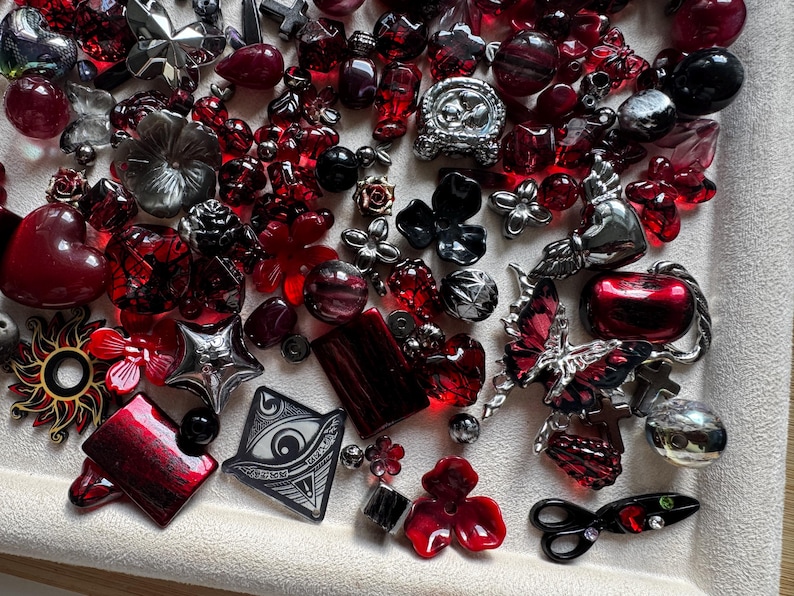 May include: A collection of assorted beads and charms in shades of red, black, and silver. The assortment includes shapes such as hearts, flowers, butterflies, crosses, stars, a sun, and an eye symbol, featuring various textures and finishes.