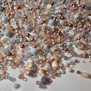 Può includere: Una collezione di perline di vari colori, rosa, pesca, argento e oro. Le perline sono di diverse forme e dimensioni, tra cui rotonde, sfaccettate e irregolari. Alcune perline hanno una lucentezza perlescente, mentre altre sono trasparenti o opache. Le perline sono sparse su una superficie bianca.