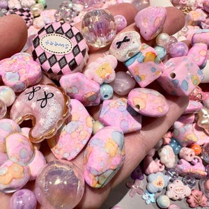 Pode incluir: Uma cole&ccedil;&atilde;o de contas e berloques rosa, brancos e iridescentes. O sortido inclui contas redondas, em forma de cora&ccedil;&atilde;o e geom&eacute;tricas, juntamente com berloques com a palavra "sweet" e outros elementos decorativos. As contas variam em tamanho e textura.