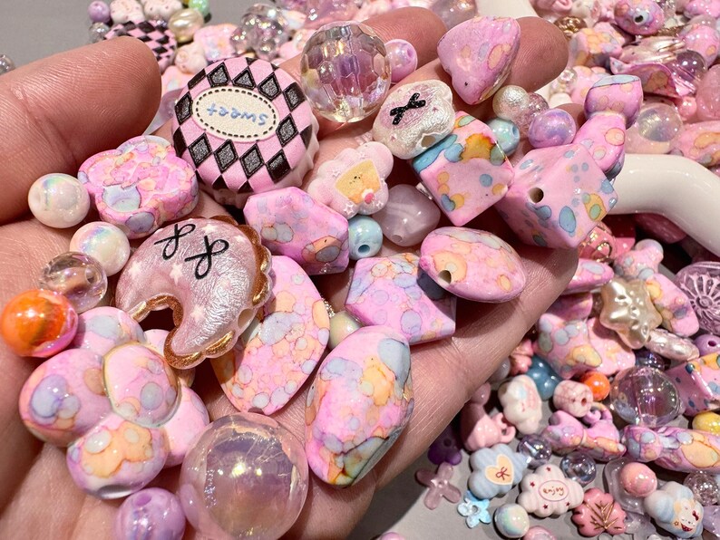 Pode incluir: Uma cole&ccedil;&atilde;o de contas e berloques rosa, brancos e iridescentes. O sortido inclui contas redondas, em forma de cora&ccedil;&atilde;o e geom&eacute;tricas, juntamente com berloques com a palavra "sweet" e outros elementos decorativos. As contas variam em tamanho e textura.
