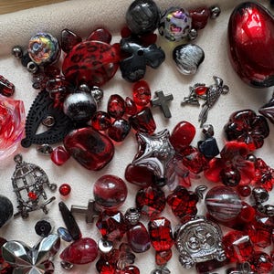 May include: A collection of assorted beads and charms in shades of red, black, and silver. The assortment includes floral shapes, crosses, stars, a bird charm, a birdcage charm, and various round, faceted, and smooth beads of different sizes.