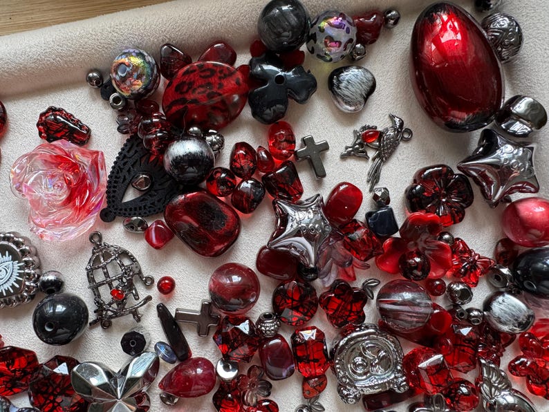 May include: A collection of assorted beads and charms in shades of red, black, and silver. The assortment includes floral shapes, crosses, stars, a bird charm, a birdcage charm, and various round, faceted, and smooth beads of different sizes.