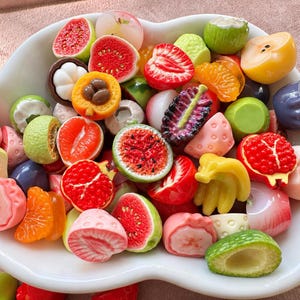 Könnte beinhalten: Eine weiße, wolkenförmige Schale gefüllt mit einer Vielzahl kleiner, bunter, fruchtförmiger Objekte. Die Sammlung enthält Miniaturdarstellungen von Erdbeeren, Wassermelonenstücken, Granatäpfeln, Bananen, Zitrussegmenten und Avocados.