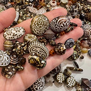 May include: A selection of artisan beads in a variety of shapes and sizes, held in a hand. The beads display intricate white and brown patterns, including floral and geometric designs. Some beads have a speckled appearance, while others are striped or solid.