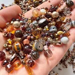 May include: A collection of beads in brown, gold, and clear hues are held in a hand. The beads vary in size and shape, including round, faceted, and irregular forms. Some beads have stripes or patterns, and others are iridescent. The beads are suitable for jewellery making.