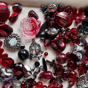 May include: A collection of assorted craft beads and charms in shades of red, black, and silver. The assortment includes translucent red rose shapes, a heart-shaped charm with an eye graphic, a birdcage charm, and various metallic cross pendants.
