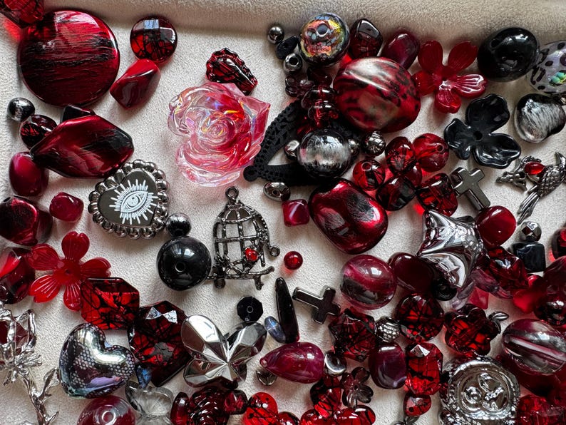 May include: A collection of assorted craft beads and charms in shades of red, black, and silver. The assortment includes translucent red rose shapes, a heart-shaped charm with an eye graphic, a birdcage charm, and various metallic cross pendants.