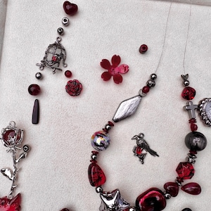 May include: A necklace featuring dark metallic and red beads, a silver bird charm, a cross, and a heart-shaped pendant with an eye design. Additional loose components include a silver rose charm, a birdcage charm, and a red flower-shaped bead.