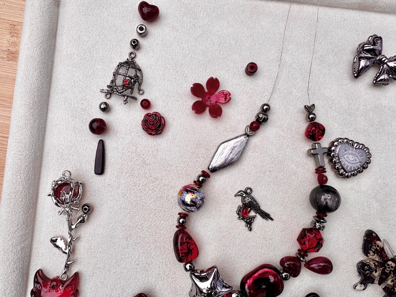 May include: A necklace featuring dark metallic and red beads, a silver bird charm, a cross, and a heart-shaped pendant with an eye design. Additional loose components include a silver rose charm, a birdcage charm, and a red flower-shaped bead.