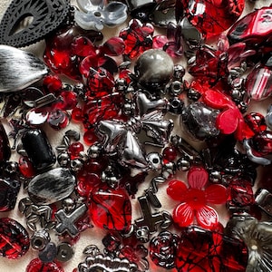 May include: A collection of assorted craft beads in shades of red, black, and silver. The mix includes various shapes such as flowers, crosses, hearts, and geometric forms with glossy, metallic, and translucent finishes for jewelry making.