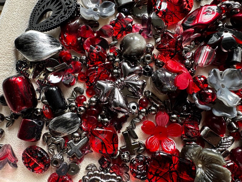 May include: A collection of assorted craft beads in shades of red, black, and silver. The mix includes various shapes such as flowers, crosses, hearts, and geometric forms with glossy, metallic, and translucent finishes for jewelry making.