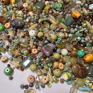 May include: A selection of beads and decorative items in green, brown, and gold hues. The assortment includes round, oval, and irregular shapes, with some featuring leaf and crystal designs. The beads vary in size and texture, creating a diverse mix.