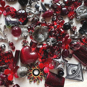 May include: A collection of assorted craft beads and charms in shades of red, black, and silver. The assortment includes heart-shaped beads, flower-shaped components, a silver rose charm, a sun-shaped pendant, and a triangular charm with an eye design.