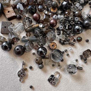 Puede incluir: Una colección de cuentas variadas en tonos gris, negro, marrón y transparente, con diversas formas que incluyen facetadas, redondas y cilíndricas. Se incluyen dos dijes de metal plateado, uno con forma de llave y otro de candado.