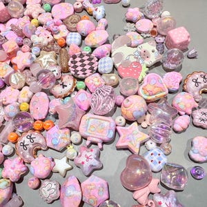 Pode incluir: Uma cole&ccedil;&atilde;o de contas e berloques rosa e pastel. O sortimento inclui estrelas, cora&ccedil;&otilde;es, flores e outras formas. Algumas contas s&atilde;o transparentes ou iridescentes, e outras t&ecirc;m padr&otilde;es ou desenhos. Os itens est&atilde;o espalhados em uma superf&iacute;cie cinza.