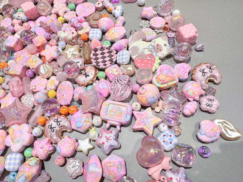 Pode incluir: Uma cole&ccedil;&atilde;o de contas e berloques rosa e pastel. O sortimento inclui estrelas, cora&ccedil;&otilde;es, flores e outras formas. Algumas contas s&atilde;o transparentes ou iridescentes, e outras t&ecirc;m padr&otilde;es ou desenhos. Os itens est&atilde;o espalhados em uma superf&iacute;cie cinza.