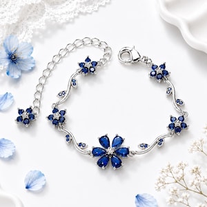 Op de afbeelding: Een zilverkleurige armband met een rankachtig ontwerp, versierd met meerdere blauwe, druppelvormige en ronde kristallen bloemen. Het sieraad is voorzien van een karabijnsluiting en een verlengketting die eindigt in een kleine blauwe kristallen bloem.
