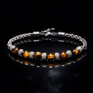 Peut inclure: Un bracelet argenté avec un motif de chaîne, orné de perles en œil de tigre et de perles argentées texturées alternées. Le bracelet est présenté sur une surface noire, reflétant la lumière.