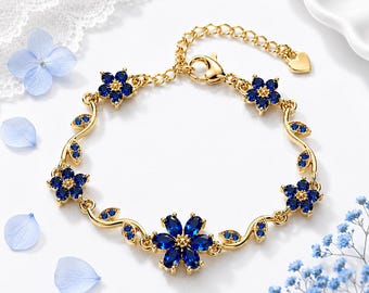 May include: A gold-tone chain bracelet featuring five flower-shaped charms and leaf-shaped accents, all set with deep blue crystals. The bracelet includes a lobster claw clasp, an adjustable chain extension, and a small heart-shaped charm at the end.