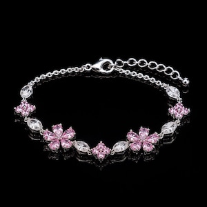 Peut inclure: Un bracelet argenté avec un motif floral délicat. Le bracelet est orné de pierres roses en forme de fleurs et de pierres claires de forme ovale. La chaîne est réglable et possède un fermoir mousqueton. Le bracelet est sur fond noir.