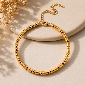 Könnte beinhalten: Ein goldfarbenes Armband mit Perlenmuster. Das Armband hat kleine, runde Perlen und zylindrische, röhrenförmige Perlen. Die verstellbare Kette und der Verschluss sind ebenfalls goldfarben. Der Schmuck wird auf einer neutralfarbenen Oberfläche präsentiert.