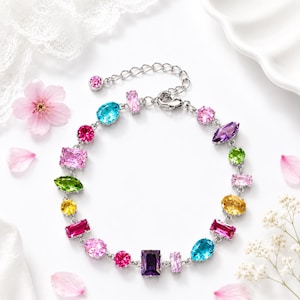 Op de afbeelding: Een zilverkleurige kettingarmband met een reeks veelkleurige, verschillend gevormde gefacetteerde kristallen, waaronder roze, blauwe, paarse, groene en gele stenen. De armband is voorzien van een karabijnsluiting en een verstelbare verlengketting.