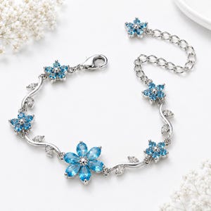 Puede incluir: Una pulsera de tono plateado que presenta una serie de flores de piedras preciosas azules y detalles de hojas de cristal transparente. El diseño incluye un cierre de mosquetón y una cadena de extensión ajustable con un delicado motivo floral.