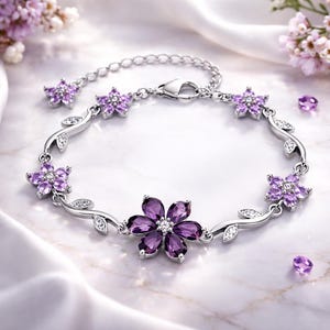 May include: A silver bracelet featuring a floral design with purple gemstone flower accents and small clear crystals. The bracelet has a delicate, feminine aesthetic, with a lobster clasp closure. The background is a marble surface.