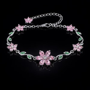 Peut inclure: Un bracelet argenté orné de pierres roses en forme de fleurs et de détails de feuilles vertes. Le bracelet, avec une chaîne délicate, est posé sur un fond noir. Le design floral est la pièce maîtresse de ce bijou.