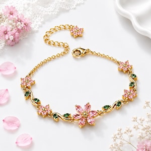 May include: A gold-tone chain bracelet featuring a series of pink crystal flowers and small green crystal leaves. The bracelet includes a lobster claw clasp, an adjustable extender chain ending in a small flower charm, and a central flower motif.