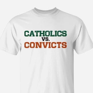 Peut inclure: Un t-shirt blanc à manches courtes avec le texte CATHOLICS VS. CONVICTS imprimé sur la poitrine. Le mot CATHOLICS est en vert foncé, l'abréviation VS. est en noir et le mot CONVICTS est en orange, le tout avec une texture vieillie.